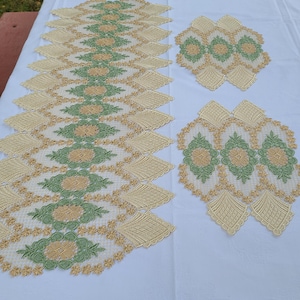 May include: Embroidered table runners and placemats in a set. The cream-coloured fabric is decorated with green and gold floral and geometric patterns. The edges of the placemats and runners are scalloped.