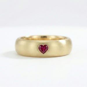 Op de afbeelding: Een gouden ring met een hartvormige robijn. De ring heeft een geborstelde afwerking en een brede band. De robijn is dieprood van kleur en is in het midden gezet. Een sieraad.