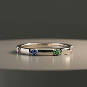 May include: A silver band ring with alternating colored gemstones. The ring features a pink, blue, green, and pink gemstone. The ring is set on a neutral surface.
