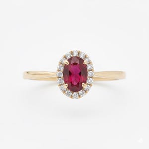 Puede incluir: Un anillo de oro con una piedra preciosa ovalada de color rojo intenso, rodeada por un halo de piedras más pequeñas y transparentes. La banda del anillo es de oro pulido y sencillo. El engaste resalta la piedra central, creando un diseño clásico y elegante.
