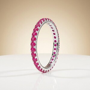 Könnte beinhalten: Ein silberner Eternity-Ring mit einer durchgehenden Reihe runder, leuchtend pinker Rubine, die in eine polierte Metallschiene gefasst sind. Der Ring steht aufrecht und zeigt das kreisförmige Design sowie die gleichmäßige Anordnung der Edelsteine.