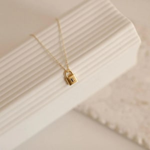 Puede incluir: Un collar dorado con un pequeño colgante en forma de candado. El colgante tiene grabada la letra "E". El collar se muestra sobre una superficie blanca texturizada. La cadena es delicada y el colgante es rectangular.