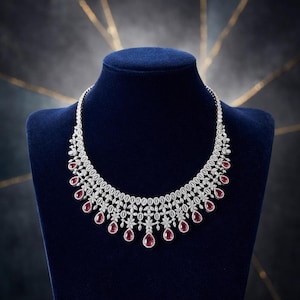 Può includere: Una collana scintillante con pietre preziose rotonde e trasparenti e pietre rosse a goccia. La collana è esposta su un busto di velluto blu, su uno sfondo scuro con accenti dorati.