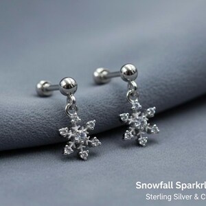 May include: Silver snowflake earrings with cubic zirconia detailing. The earrings feature a post and ball fastening. The image includes the text "Snowfall Sparkle Earrings".