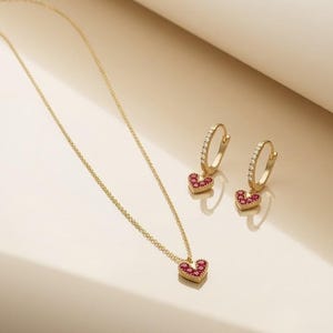 Puede incluir: Un collar de oro y un par de pendientes de aro a juego. El collar presenta un pequeño colgante en forma de corazón con piedras rojas. Cada aro tiene una fila de piedras transparentes y un dije colgante en forma de corazón con piedras rojas.