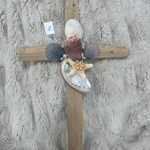 May include: A wooden cross decorated with seashells, a starfish, and faux pearls. The cross is made of light brown wood, with a vertical and horizontal beam. The seashells are various colors, including white, brown, and gray.