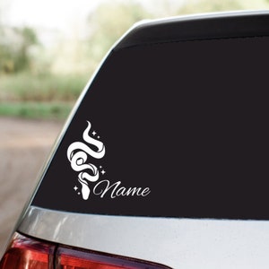May include: White vinyl decal of a stylized snake with stars on a black car window. The decal includes the word "Name" below the snake.