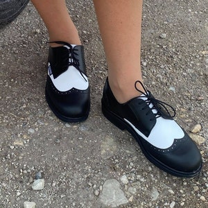 May include: Black and white leather oxford shoes with a wingtip design and laces. The shoes have a white toe cap and a black heel.