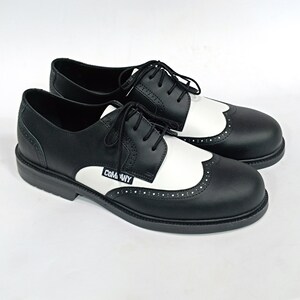 May include: Black and white leather shoes with a wingtip design and a black rubber sole. The shoes have a white leather panel on the side with the word "COMME des GARCONS" printed in black.