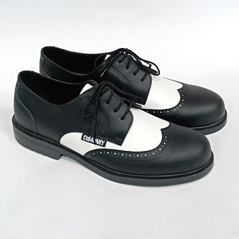 May include: Black and white leather shoes with a wingtip design and a black rubber sole. The shoes have a white leather panel on the side with the word "COMME des GARCONS" printed in black.