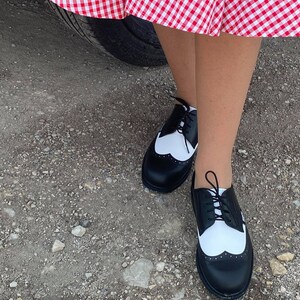 May include: Black and white leather oxford shoes with a wingtip design and laces. The shoes are worn by a person standing on a gravel surface.