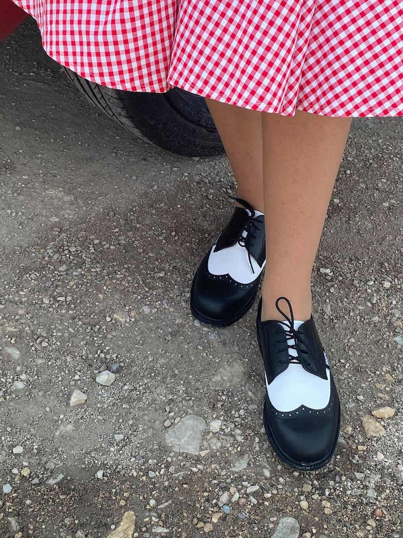 May include: Black and white leather oxford shoes with a wingtip design and laces. The shoes are worn by a person standing on a gravel surface.