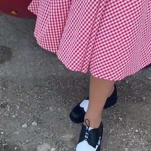 May include: Black and white two-tone oxford shoes with a heart-shaped detail on the toe. The shoes are worn with a red and white gingham skirt.