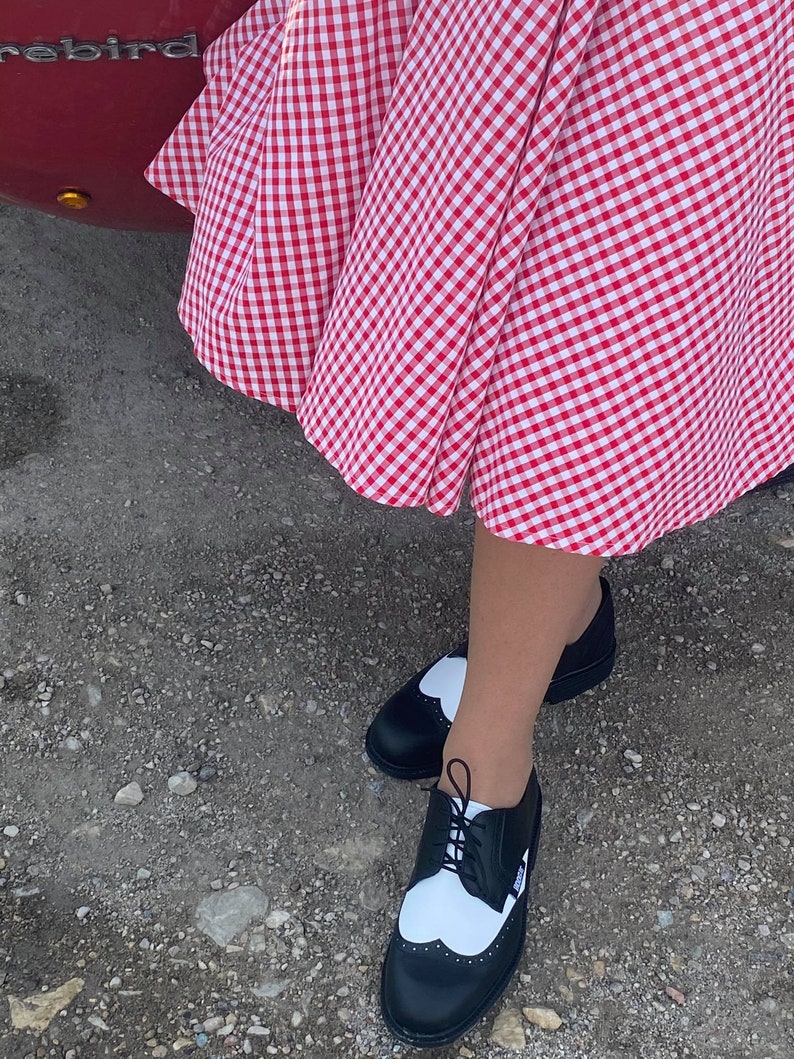 May include: Black and white two-tone oxford shoes with a heart-shaped detail on the toe. The shoes are worn with a red and white gingham skirt.