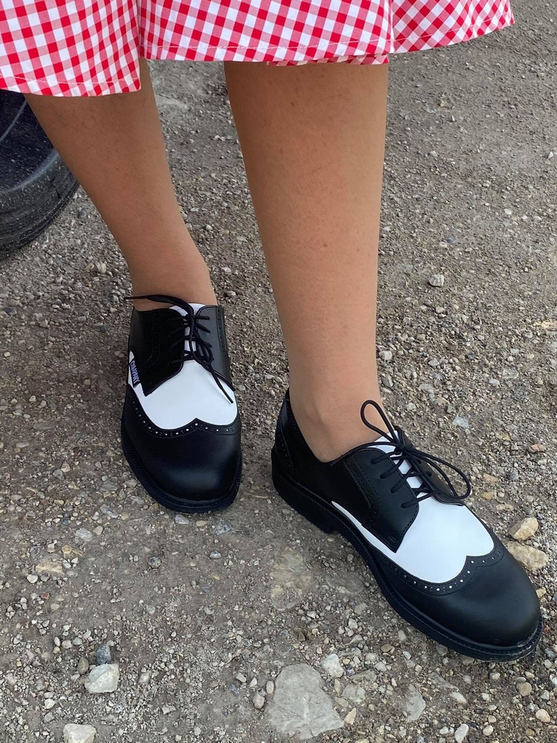 May include: Black and white leather oxford shoes with a wingtip design and laces. The shoes have a white toe cap and a black heel.