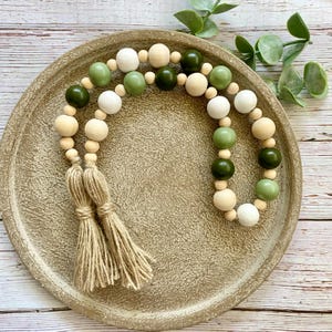 May include: A wooden bead garland with green and white beads and natural jute tassels. The garland is displayed on a brown plate.