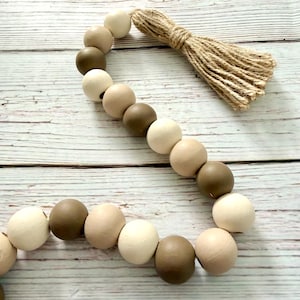 May include: A decorative wooden bead garland in shades of cream, tan, and brown. The beads are strung together and accented with a natural jute tassel. The garland is arranged on a light wood surface.