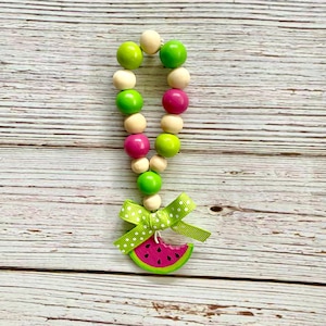 May include: A wooden bead garland with green, pink, and natural wood beads. The garland has a watermelon charm attached to the bottom.