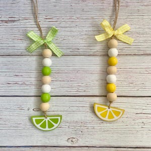 May include: Two beaded ornaments with a citrus fruit theme. One features a lime slice and green and white beads, the other a lemon slice with yellow and white beads. Both have a gingham bow and a twine loop for hanging.