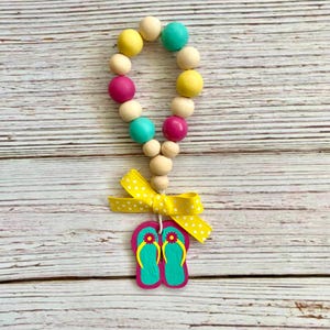 May include: A wooden bead garland with yellow, pink, and blue beads. A yellow bow with white polka dots is attached to the bottom of the garland. A pair of turquoise flip-flops with pink accents are attached to the bow.