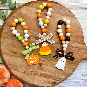 May include: Three Halloween-themed beaded garlands with wooden beads in orange, white, black, and green. Each garland has a different Halloween-themed charm: a pumpkin, a candy corn, and a ghost. The charms are attached to a ribbon with a Halloween-themed print.