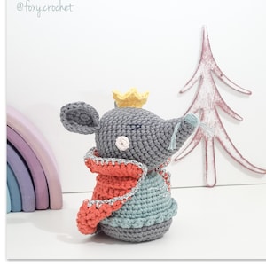 May include: A crocheted gray mouse wearing a yellow crown, a red and silver cape, and a blue and gray outfit. The mouse has closed eyes and a light blue whisker. The background includes a wooden rainbow and a pink tree cutout. The text "foxy.crochet" is in the top left corner.