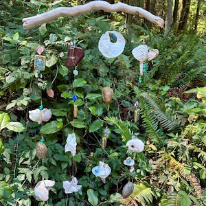 May include: A nature-inspired mobile with various ceramic and shell ornaments hanging from a driftwood branch. The mobile features a variety of colours, including white, brown, blue, and green.