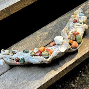 May include: A piece of driftwood decorated with colorful stones and seashells. The driftwood is light brown and has a natural, weathered look. The stones are various shades of red, green, and brown. The seashells are white and have a smooth, polished surface.