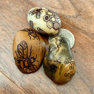 May include: Three painted stones with bee and honeycomb designs. The stones are brown, gray, and white with black ink drawings.