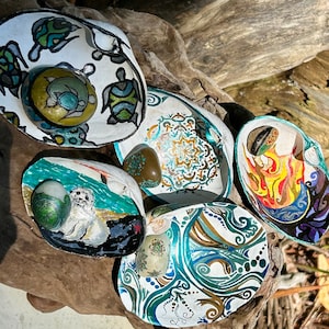 May include: Five seashells painted with different designs. Each shell has a smooth, white interior and a colourful exterior. The designs include a mandala, a sea turtle, a seal, a flame, and a swirling pattern. Each shell has a smooth, round river rock inside.