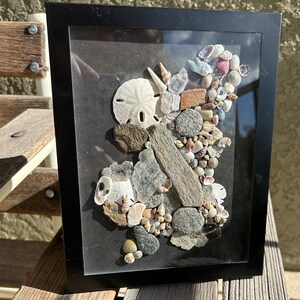 May include: A framed artwork featuring a seashell and stone mosaic in a black frame. The mosaic is made up of various seashells, stones, and driftwood, creating a natural and organic design.