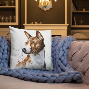 May include: A watercolor painting of a brown and white Basenji dog on a white pillow with colorful splatter paint. The dog is looking to the left of the image.
