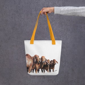 Op de afbeelding: Een witte tote bag met oranje handvatten heeft een aquarel schilderij van vijf bruine koeien. De koeien staan in een rij en kijken naar de kijker.