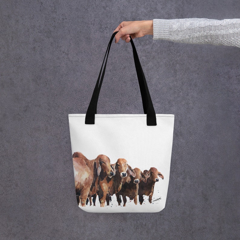 Op de afbeelding: Een witte tote bag met zwarte handvatten heeft een aquarel schilderij van vijf bruine koeien die naar de camera kijken.