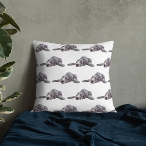 Peut inclure: Coussin carré blanc avec un motif répété de chien de style aquarelle. Les chiens sont représentés couchés, la tête posée. Les chiens sont gris et blancs, sur un fond blanc. Le coussin est sur une couverture bleu foncé.