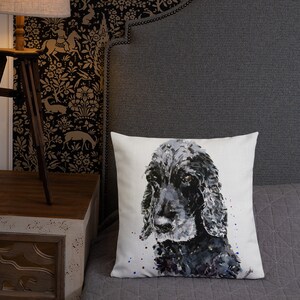 Può includere: Un cuscino decorativo con un dipinto ad acquerello di un cane Cocker Spaniel nero. Il pelo del cane è raffigurato in tonalità di nero, grigio e bianco, con schizzi di vernice colorati. Il cuscino è su un letto con una trapunta grigia.