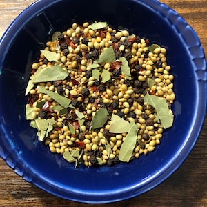 May include: A blue bowl filled with a variety of spices, including bay leaves, peppercorns, coriander seeds, and red pepper flakes.