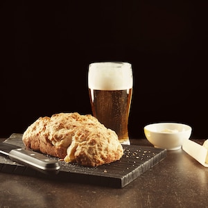 May include: A loaf of bread on a black cutting board with a knife, a glass of beer, a small white bowl, and a piece of butter on a white napkin.