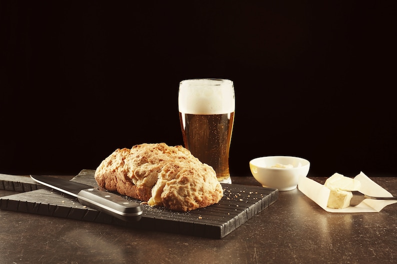 May include: A loaf of bread on a black chopping board with a knife, a glass of beer, a small white bowl, and a piece of butter on a white napkin.