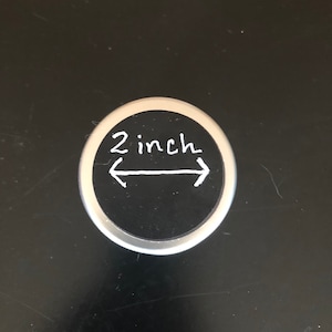 May include: A black and silver round sticker with white text that reads "2 inch" and two white arrows pointing in opposite directions.