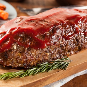 May include: A cooked meatloaf, glazed with a red sauce, sits on a wooden cutting board. Fresh rosemary sprigs are placed around the meatloaf. Sliced carrots are visible in the background, suggesting a complete meal.