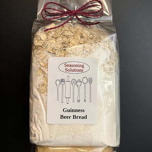 May include: A clear plastic bag with a red ribbon tied around it. The bag contains a mix of dry ingredients for making Guinness Beer Bread. The label on the bag reads "Seasoning Solutions" and "Guinness Beer Bread". The net weight is 19 oz (532g).