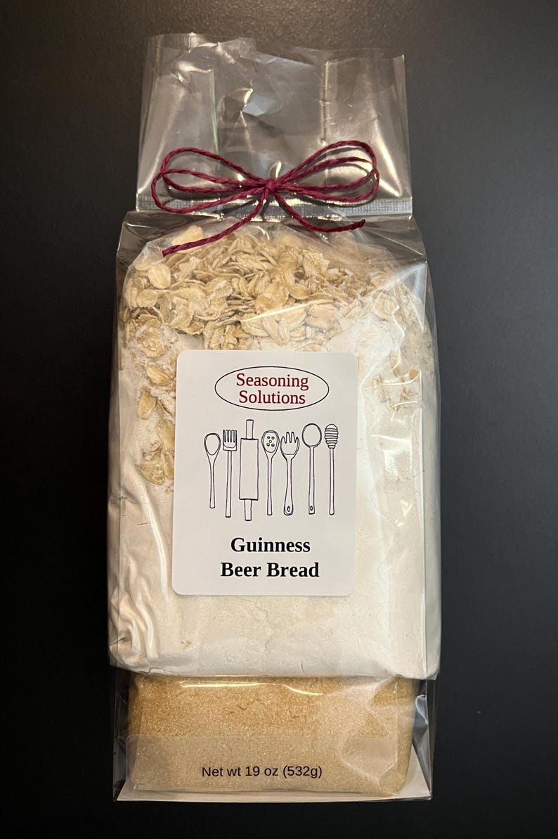May include: A clear plastic bag with a red ribbon tied around it. The bag contains a mix of dry ingredients for making Guinness Beer Bread. The label on the bag reads "Seasoning Solutions" and "Guinness Beer Bread". The net weight is 19 oz (532g).