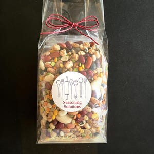 May include: A clear plastic bag tied with a red ribbon contains a mixture of dried beans, lentils, and peas. The bag has a white label with the text "Seasoning Solutions" and a drawing of kitchen utensils. The label also states "Net Wt 16 oz (454g)".