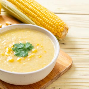 May include: A bowl of creamy corn soup garnished with a sprig of cilantro, alongside fresh corn on the cob and loose kernels. The soup is served on a wooden cutting board, with a light-colored wooden background.