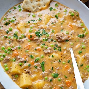 May include: A bowl of creamy soup with potatoes, peas, carrots, and ground meat, garnished with fresh parsley. A square cracker rests on the soup's edge, and a silver spoon is in the soup. The soup is in a white bowl.