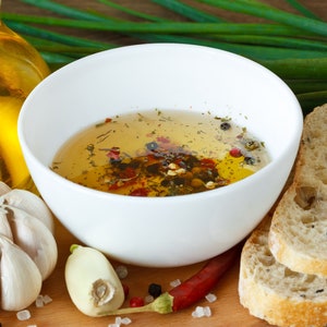 May include: A white bowl filled with a clear olive oil dressing with herbs and spices. The bowl is on a wooden surface with garlic cloves, a sprig of green onions, and a red chili pepper.