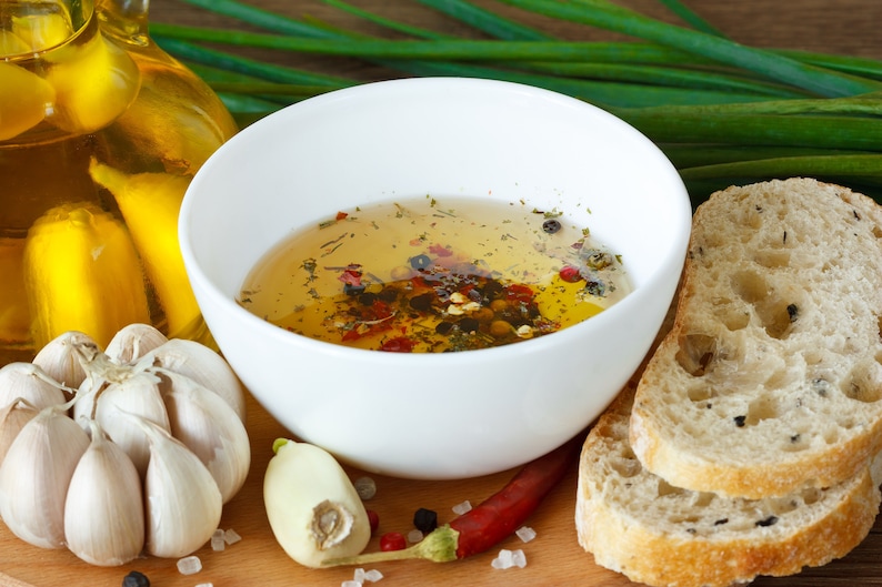 May include: A white bowl filled with a clear olive oil dressing with herbs and spices. The bowl is on a wooden surface with garlic cloves, a sprig of green onions, and a red chili pepper.