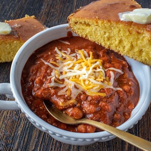 May include: A white bowl filled with chili topped with shredded cheese and a golden spoon. Two slices of cornbread are on the side, one with a pat of butter.