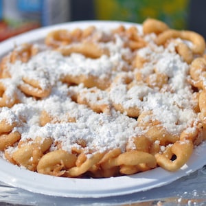 May include: A white paper plate with a large serving of funnel cake covered in powdered sugar.